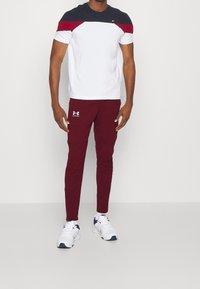 Herrarnas sportoutfit: vit och marinblå kortärmad skjorta med röda detaljer; vinröda tajta byxor; vita sneakers med marinblå detaljer.