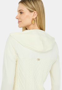 Cardigan bianco con cappuccio, caratterizzato da un motivo a treccia testurizzata sulla schiena e un discreto dettaglio in metallo dorato vicino al collo.
