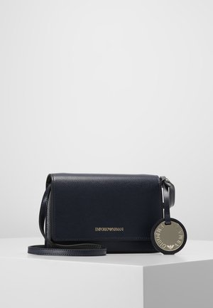Granatowa skórzana torba typu crossbody Emporio Armani z złotym logo i okrągłą metalową metką z logo na białej powierzchni wystawowej.