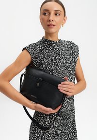Borsa a tracolla nera in pelle con finitura testurizzata, dotata di chiusura a bottone automatico e tracolla regolabile. Esposta con un abito a fantasia.