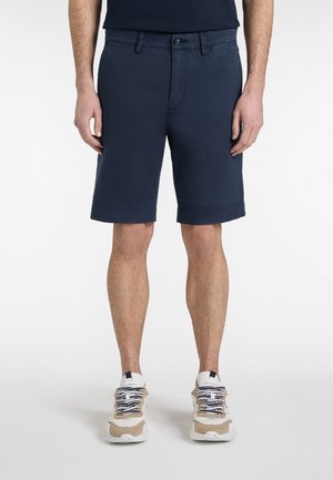 Moški, oblečen v temno mornarske kratke hlače in bele superge, stoji z rokami sproščeno ob telesu na enobarvnem belem ozadju.