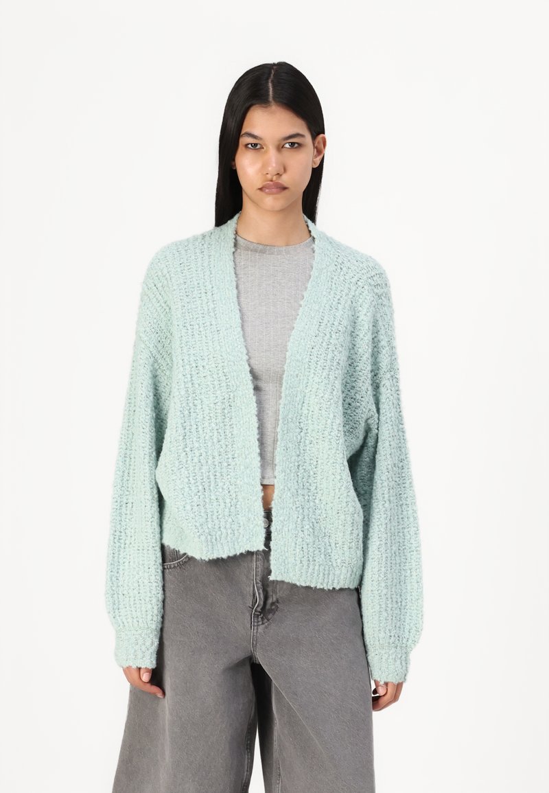 Ljusblå stickad cardigan med lös passform, ribbad textur och vida ärmar, i kombination med en grå crop top och grå vida byxor.