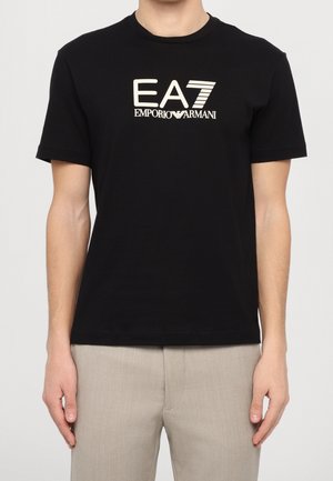 Person trägt ein schwarzes T-Shirt mit kurzen Ärmeln und weißem "EA7 Emporio Armani" Logo sowie beigefarbene Hosen, dargestellt vom Hals bis zur Hüfte.