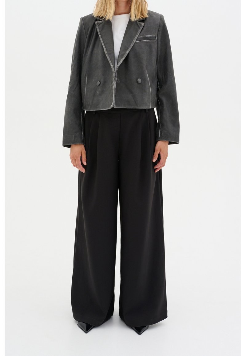 Femme portant une veste courte à double boutonnage gris foncé, un top blanc, un pantalon large noir et des chaussures noires pointues sur un fond uni.