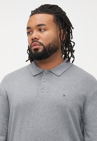 Grå polo skjorte laget av teksturert stoff, med en brettet krage, tre knapper, og en liten logo med røde, hvite og blå detaljer.