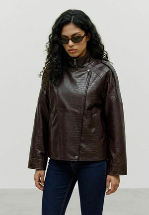 Femme aux longs cheveux bouclés, portant des lunettes de soleil foncées, une veste en cuir texturée marron foncé et un jean bleu foncé sur un fond uni.