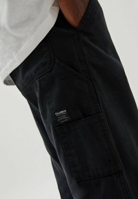 Pantalones cargo negros con una tela texturada, que presentan un bolsillo lateral y una etiqueta de marca. Están combinados con una camiseta blanca a la cintura.