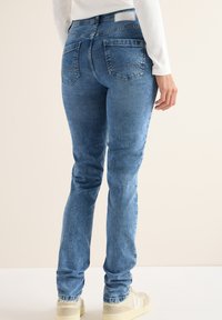 Jeans de denim azul claro con corte slim, que cuentan con bolsillos traseros y sutiles detalles de costura. Combinados con zapatillas de color claro.