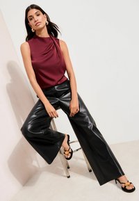 Bordeauxrotes ärmelloses Satin-Oberteil mit drapiertem Design, kombiniert mit schwarzen, weit geschnittenen Hosen aus Kunstleder und schwarzen Sandalen mit runden Akzenten.