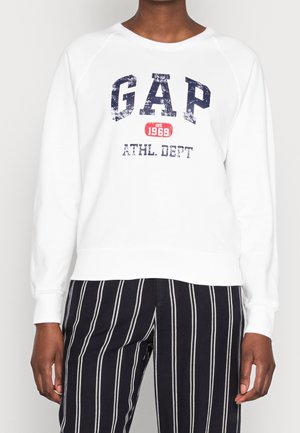 Persona che indossa una felpa bianca con la scritta “GAP Est. 1969 ATHL. DEPT” e pantaloni neri con strisce verticali bianche, in piedi contro uno sfondo semplice.