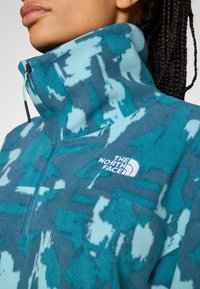 Fleecejacke in blaugrün mit abstraktem Muster, hohem Kragen und Halbverschluss. Mit weißem Logo auf der Brust. Weicher Stoff.