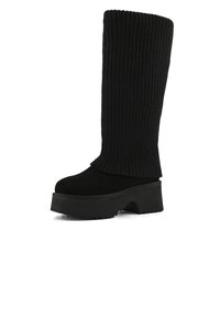 Černé vysoké boty do kolen s ribbed textilním svrškem, surovým povrchem, silnou platformou a zaoblenou špičkou. Žádné viditelné akcenty nebo kovové prvky.