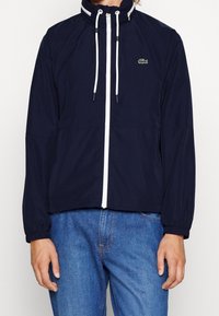 Marinblå vindjacka med hög krage, vit dragkedja och justerbara snoddar. Har en liten grön logotyp på bröstet.