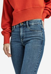 Blå denimjeans med en tettsittende design, med knappelukking og en merketag på livet. Kombinert med en kort rød hettegenser.