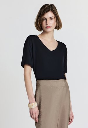 Donna con capelli corti castani, che indossa un top nero a scollo a V, una gonna beige e un bracciale spesso marmorizzato, in piedi davanti a uno sfondo bianco semplice.