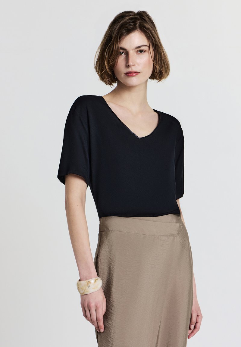 Donna con capelli corti castani, che indossa un top nero a scollo a V, una gonna beige e un bracciale spesso marmorizzato, in piedi davanti a uno sfondo bianco semplice.