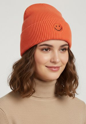 Orange knitted beanie with ribbed texture and a small embroidered smiley face on the front. Worn with a beige ribbed turtleneck sweater.
