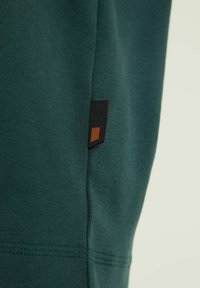 Grön bomullströja med en svart och brun märkesetikett på sidosen. Slät textur, figurnära passform och rena sömmar.