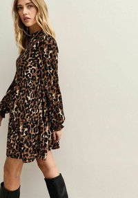 Vestido de estampado de leopardo con mangas largas de volantes, un escote alto y una silueta en capas. Confeccionado en un tejido ligero y texturizado en tonos marrón y negro.