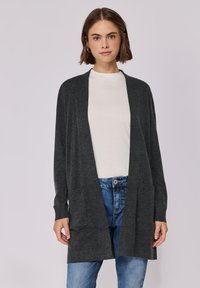Kohlenstofffarbener Cardigan mit offenem Frontbereich, langen Ärmeln, zwei Fronttaschen und einem weichen, strukturierten Strickdesign. Getragen über einem weißen Oberteil und Jeans.