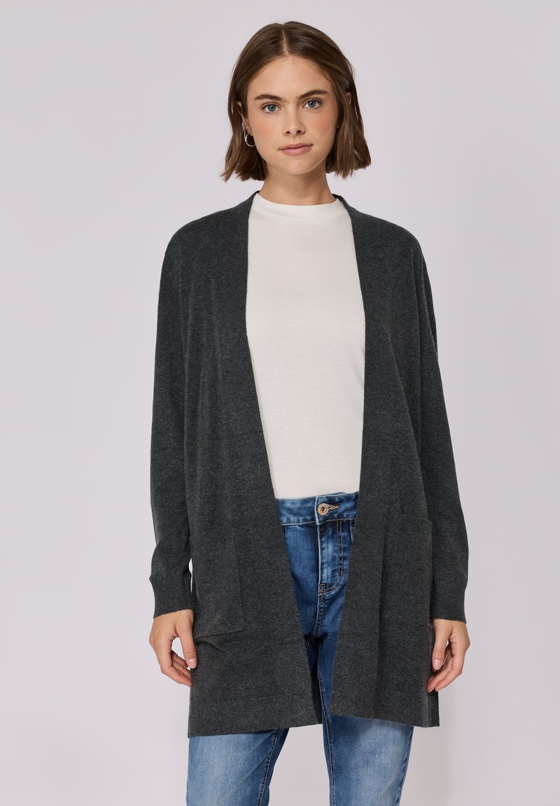 Kohlenstofffarbener Cardigan mit offenem Frontbereich, langen Ärmeln, zwei Fronttaschen und einem weichen, strukturierten Strickdesign. Getragen über einem weißen Oberteil und Jeans.