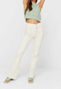 White flared jeans with a smooth texture and front pockets, paired with a light green knitted cropped tank top and matching green flip-flops.