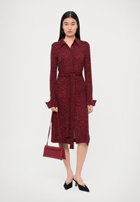 Rochie roșie cu imprimeu leopard, cu un brâu, mâneci lungi și închidere cu nasturi; asortată cu pantofii cu vârf ascuțit negri și o geantă mică roșie.