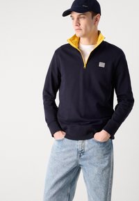 Un hanorac bleumarin cu fermoar până la jumătate, având o guler galben, dotat cu o emblemă pe partea stângă a pieptului. Purtați cu blugi din denim de culoare albastră deschisă.