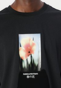 Sort bomulds T-shirt med et grafisk motiv af en pink blomst i et rektangulært design, med japansk tekst og hvidt broderet skrift nedenfor.