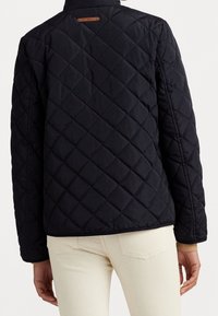 Černá prošívaná bunda s vysokým límcem, zdobená diamantovým prošitím a hnědým koženým štítkem na zadní straně. Hladká textura a přiléhavý střih.