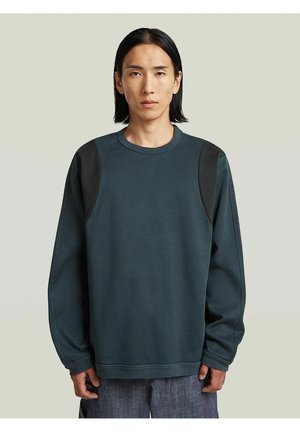 Uomo con capelli neri lisci che indossa una felpa a maniche lunghe verde acqua scuro con toppe nere sulle spalle, su uno sfondo chiaro e uniforme.