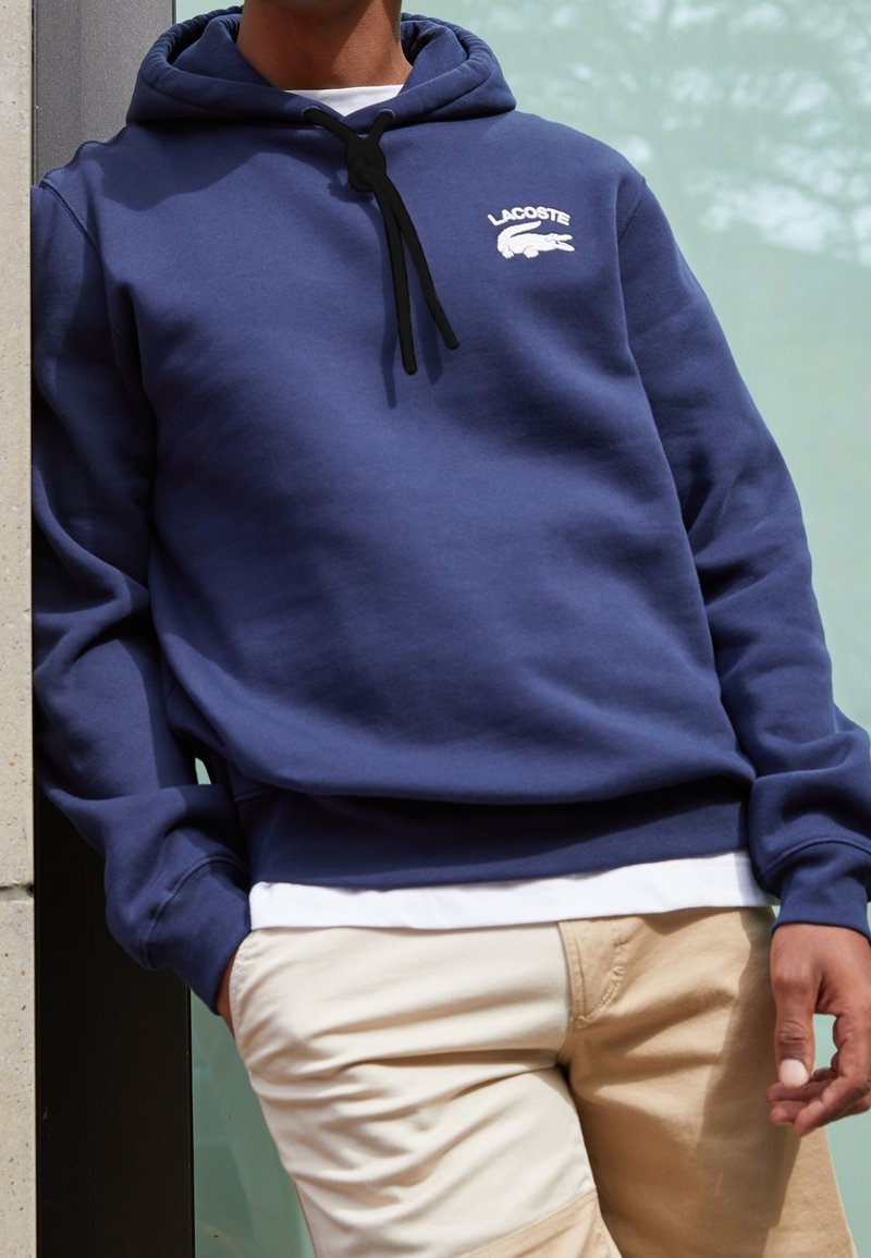 Personne portant un sweat à capuche Lacoste bleu marine avec le logo sur la poitrine, un sous-pull blanc et un pantalon beige, debout devant un mur en verre et béton.