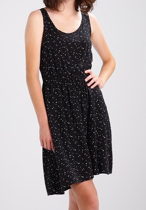 Ärmelloses schwarzes Kleid mit weißen Polka-Dots, taillierter Passform und knielangem Saum, getragen von einer Person mit einer Hand in der Hüfte.