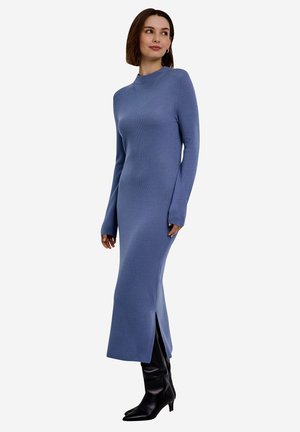 Femme portant une robe en tricot bleue à manches longues et col haut avec une fente latérale, ainsi que des bottes noires à talons mi-mollet, debout et regardant sur le côté.