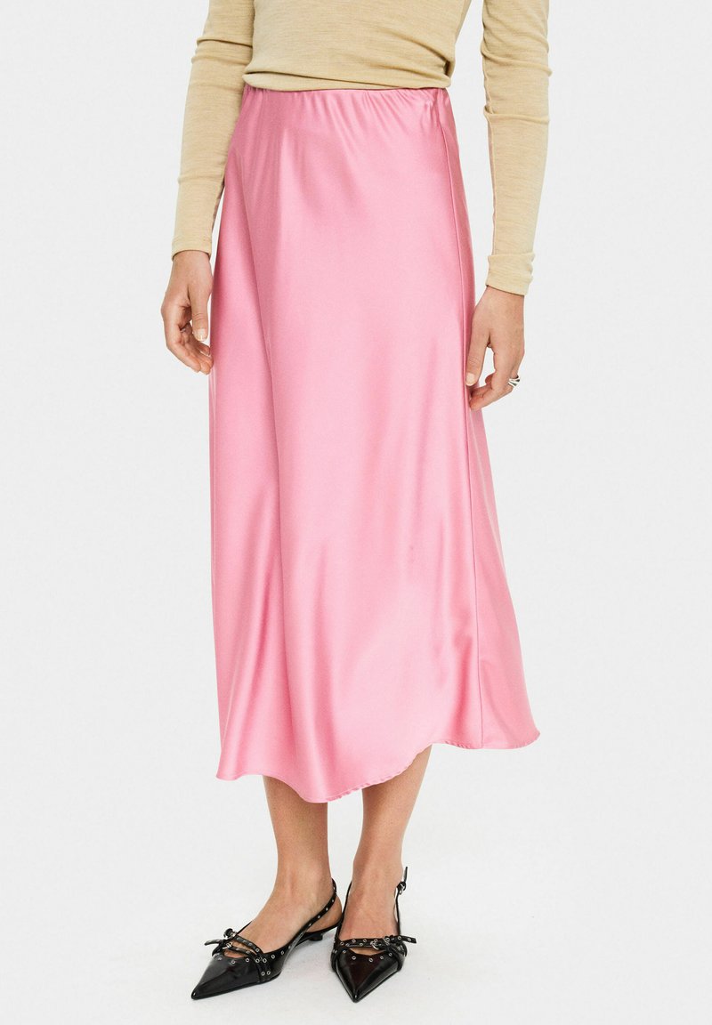 Roze satijnen midi-jurk met een gladde textuur en elastische tailleband, gecombineerd met zwarte schoenen met een spitse neus en decoratieve accenten.