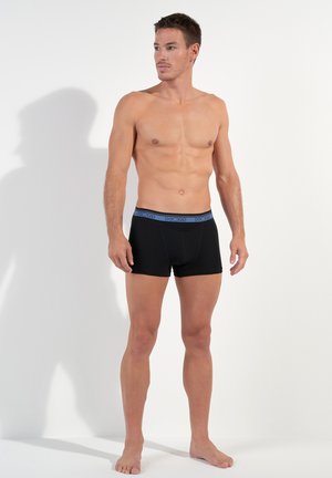 Zwarte boxershorts met een blauwe tailleband, van katoen, aansluitend ontwerp, zij aanzicht van model, blote voeten, staand tegen een neutrale achtergrond.