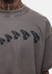 Grijze short-sleeve t-shirt van zachte stof met een zwart vlinderontwerp dat over de borst is gedrukt. Gestikte halslijn en een relaxte pasvorm.