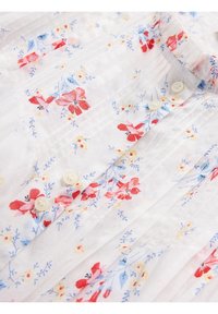 Tissu blanc avec un motif floral rouge, bleu et jaune, avec de petits boutons et de fines plis verticaux près du col et à l'avant.