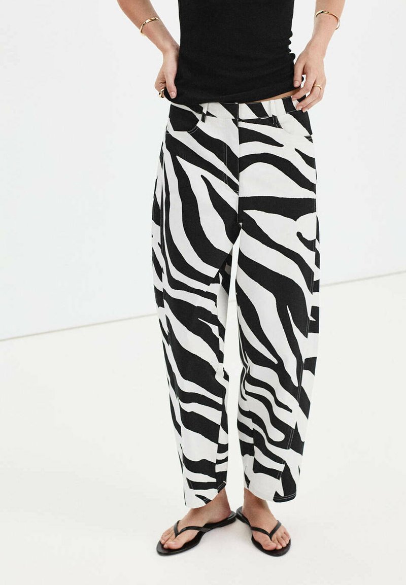 Personne portant un pantalon large imprimé zèbre noir et blanc, un haut noir sans manches, des tongs et des bracelets en or aux poignets.