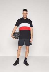 Schwarzes Poloshirt mit weißen und roten horizontalen Streifen, kombiniert mit dunklen Shorts und schwarzen Turnschuhen. Das Modell hält einen Basketball.