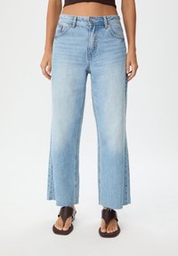 Světle modré široké džíny vyrobené z denimu, s vysokým pasem, syrovým lemem a bočními rozparky. V kombinaci s hnědými plochými sandály.