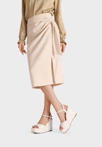 Gonna avvolgente beige con vita legata e spacco frontale, realizzata in un tessuto morbido, abbinata a sandali con zeppa bianchi con cinturini alla caviglia.