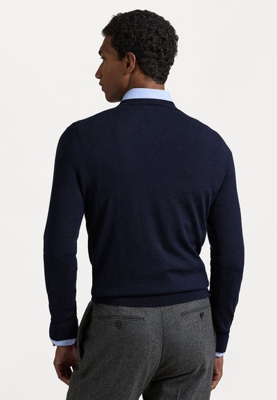 Pull bleu marine avec une finition texturée et des manches longues, associé à un pantalon gris comportant deux boutons à l'arrière et une chemise bleu clair en dessous.