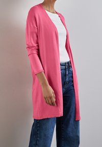 Dlouhý růžový kardigan s volným střihem, žebrovanými manžetami a otevřeným předním dílem. Nositý přes bílé tričko a spárovaný s modrými džíny. Hladká textura.