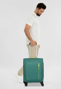 Valise roulante teal avec une surface texturée, dotée de roues noires, d'un accent de fermeture éclair jaune et d'une poignée rétractable.