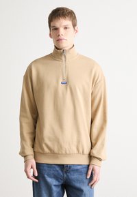 Felpa beige con zip a un quarto, colletto alto, polsini e vita a costine. Presenta un piccolo logo blu sul petto. Tessuto morbido.