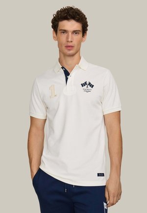 Hombre con polo blanco de manga corta con ribete azul marino y logo de banderas cruzadas, de pie frente a un fondo beige liso, con las manos en los bolsillos.