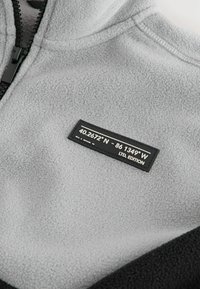 Grå fleece stof med en sort sektion, der har en sort label med koordinater og teksten "LTD. EDITION". Lynlås lukning foran.