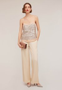 Pailletten-Camisole in Champagner mit dünnen Trägern, kombiniert mit weiten cremefarbenen Hosen und einer braunen Clutch. Hat eine glatte, glänzende Textur.