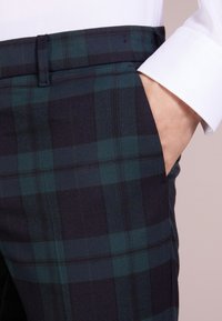 Pantalones de cuadros en verde oscuro y azul marino, con corte recto, bolsillo lateral y textura de tela suave. Detalles en la cinturilla a medida.